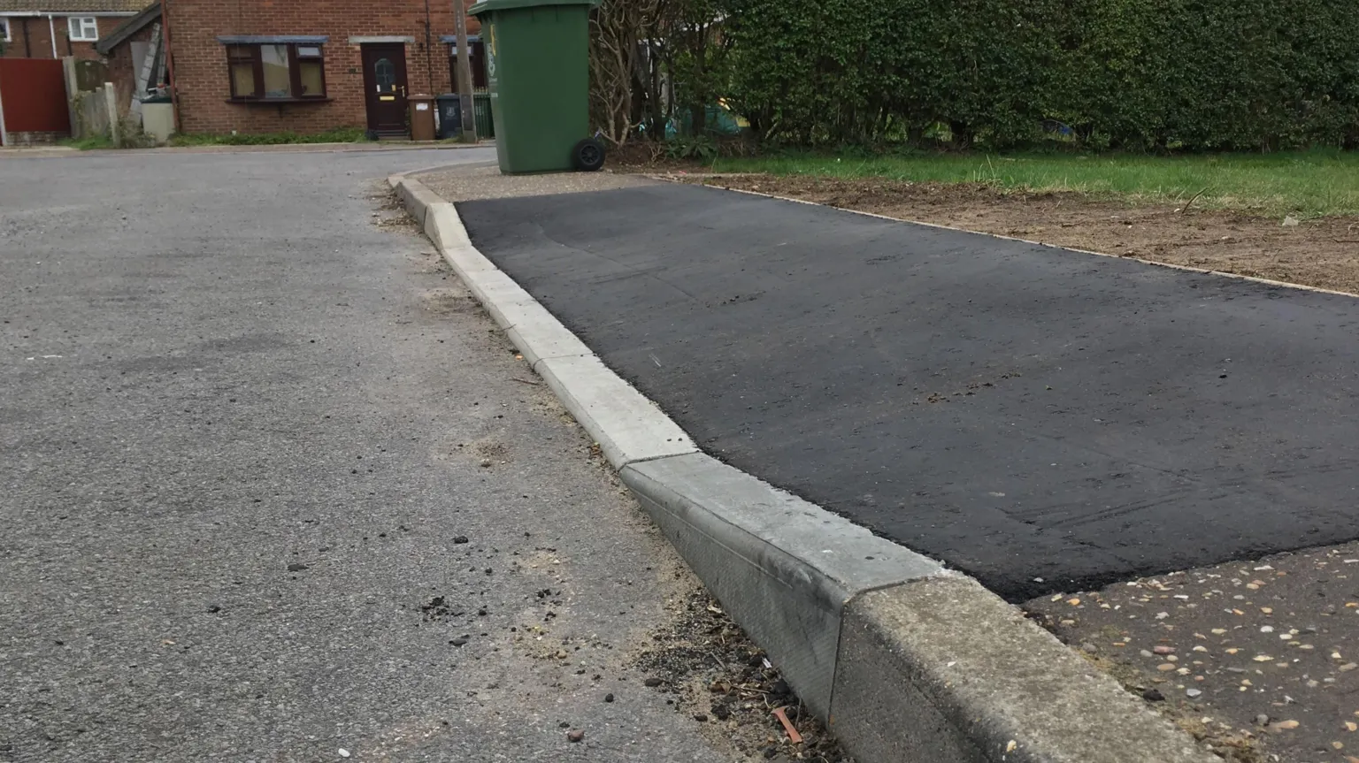A dropped kerb. The tarmac is dark grey. A green wheelie bin can be seen in the distance as well as a house.