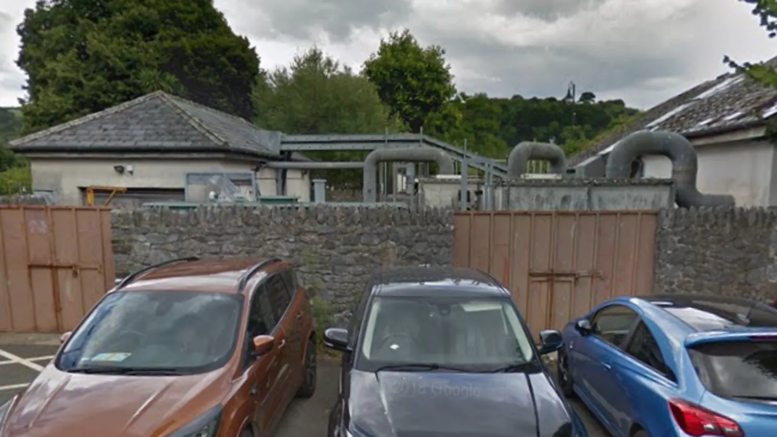 Google A small car park with several parked cars sits in front of a stone wall and a low industrial-looking site with pipes and buildings, likely part of a water or sewage facility, surrounded by trees.