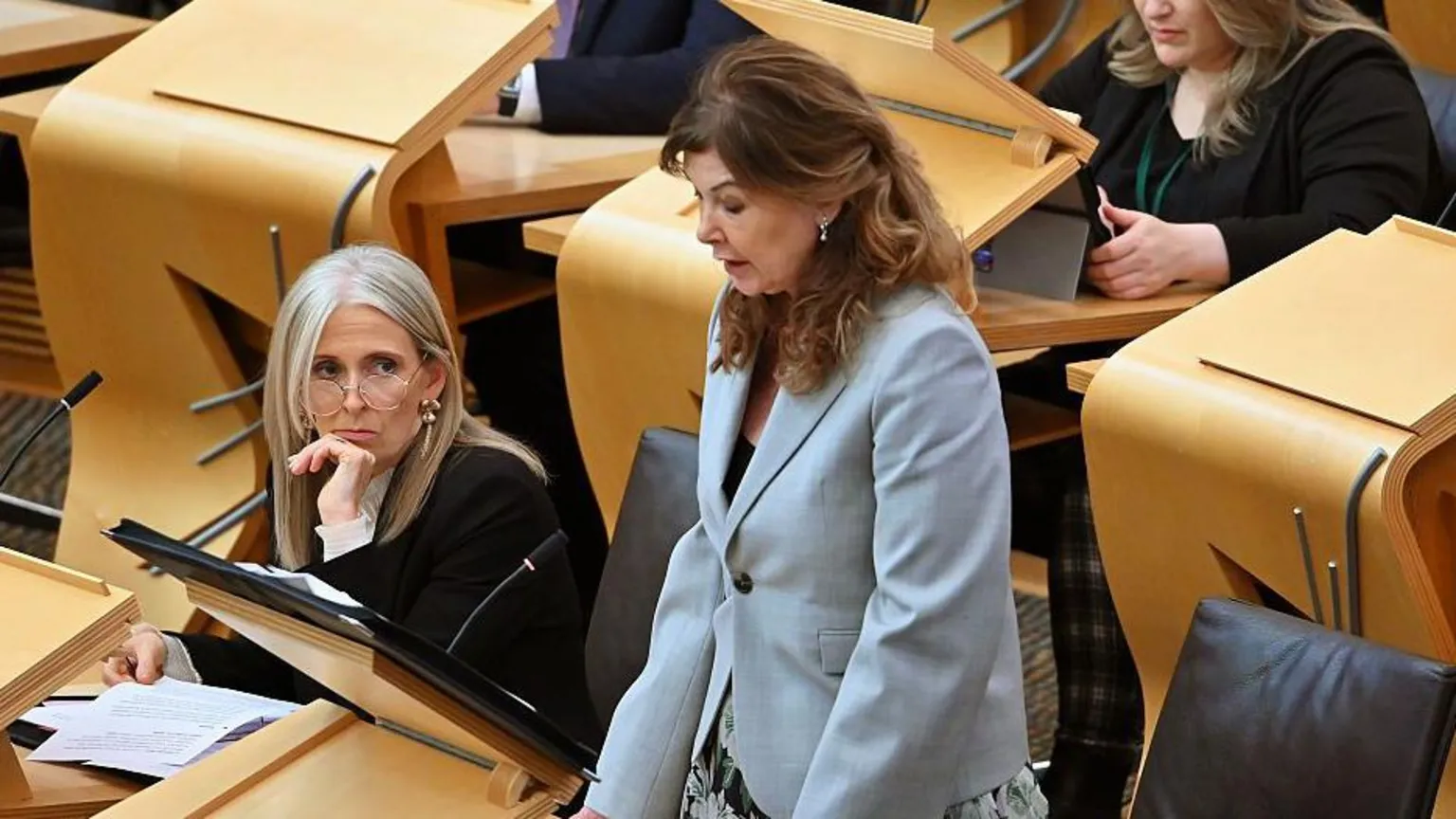 Ruth Charteris KC and Dorothy Bain KC in the Holyrood chamber. Charteris has blonde hair and is wearing a black blazer, white blouse and glasses. She is sitting down. Bain is standing. She has brown hair and is wearing a blue jacket and floral skirt.