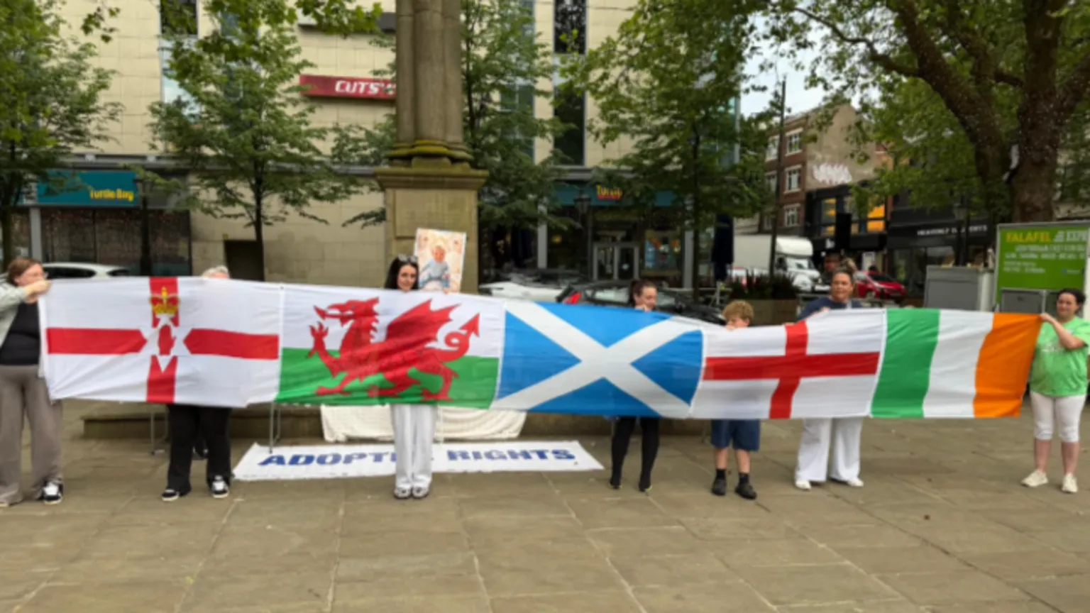 People at a vigil for murdered baby Preston Davey in Preston's Flag Market holding up flags. People are starting to arrive at the vigil to remember the 13-month-old. An image of Preston can be seen in the background and a sign saying 'Adoptee Rights'.