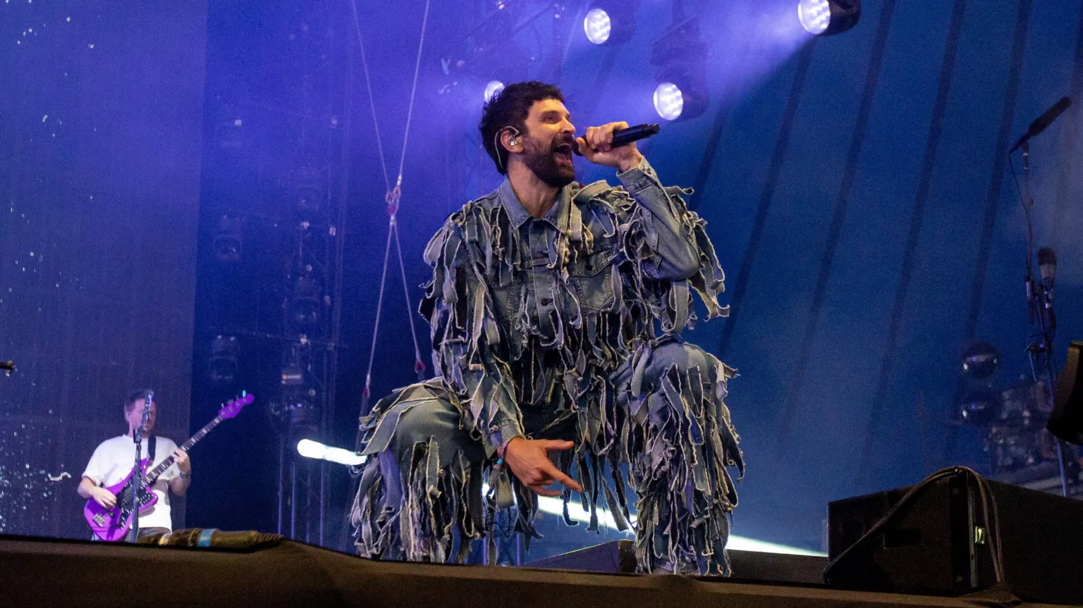 Serge Pizzorno on stage with Kasabian. He is crouched down, wearing an outfit that appears to be in tatters. A guitarist is visible in the background.