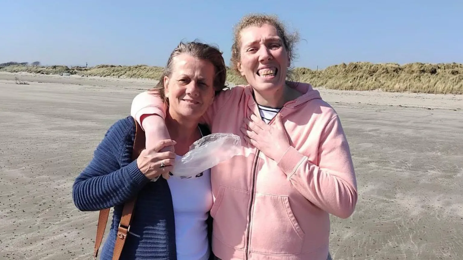 Two women standing with their arms around each other on a beach. The woman on the left is wearing a white top and a navy jacket. The woman on the right is wearing a pink zip-up hoodie. They are both smiling at the camera.
