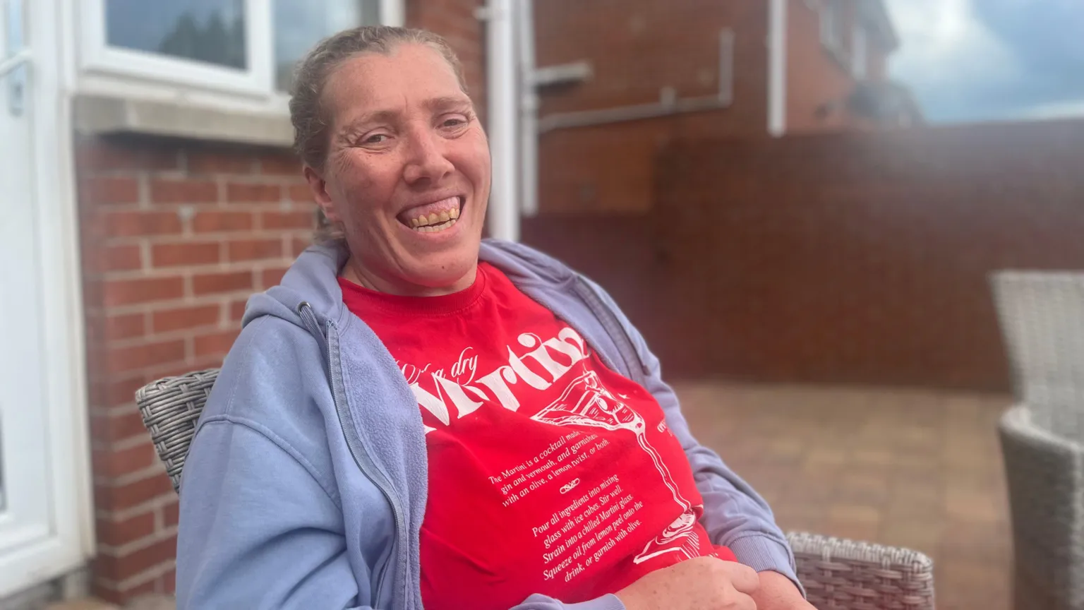 Laura is picture looking toward the camera and smiling. She's wearing a bright red t-shirt with white writing on it and a grey zip-up hoodie. She's sitting on a wicker chair outside a red brick home.