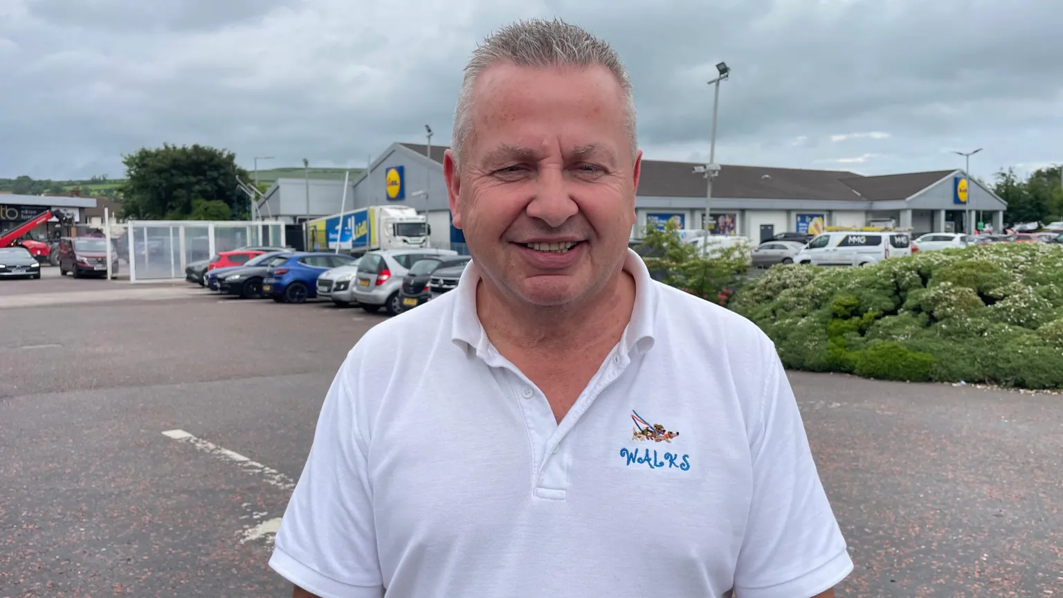 A man, with short grey hair, wearing a white t-shirt. He is standing in a carpark with a Lidl store behind him. 