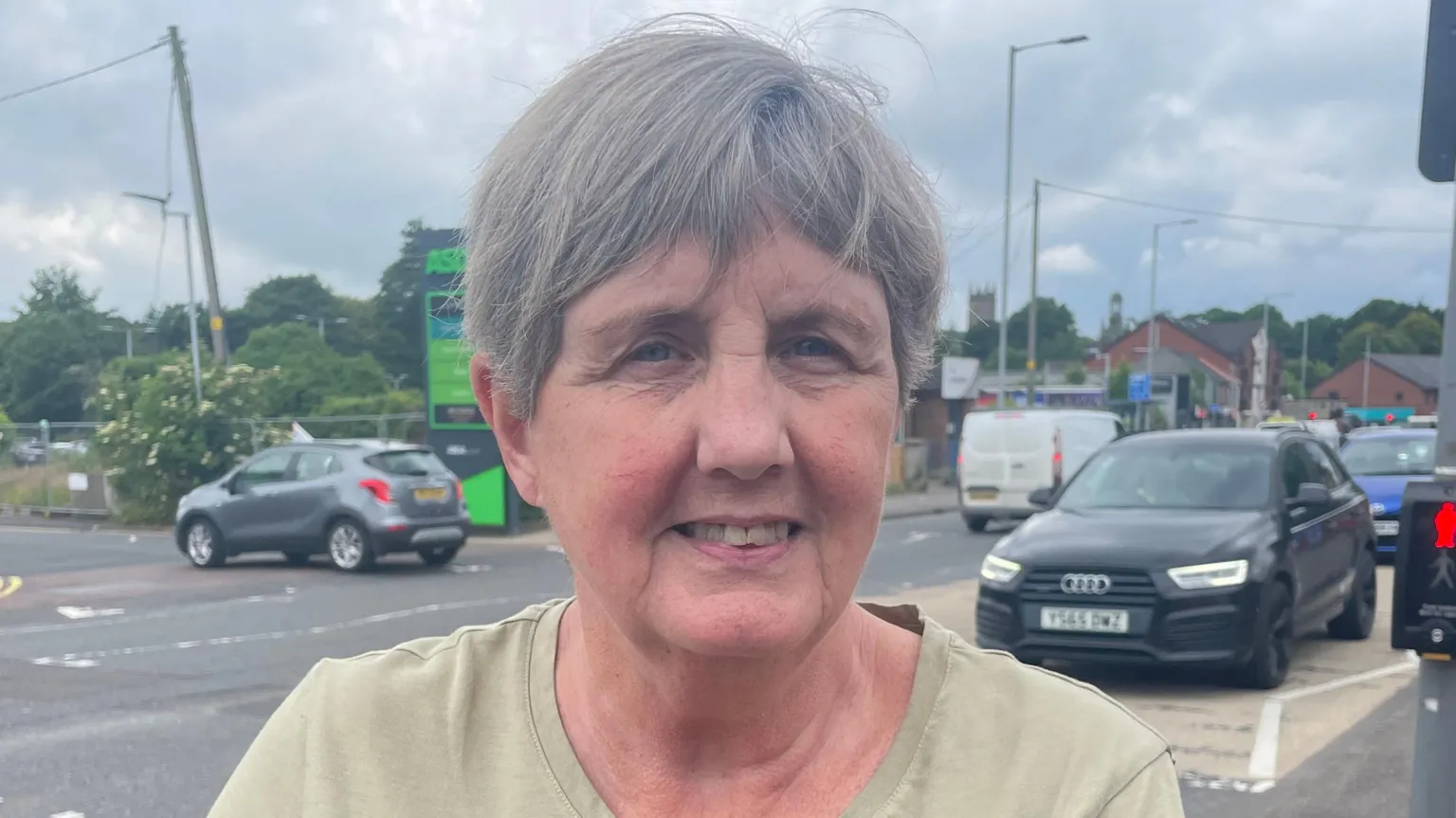 A woman, with short grey hair, wearing a light green t-shirt. She is standing on the side of the road. 