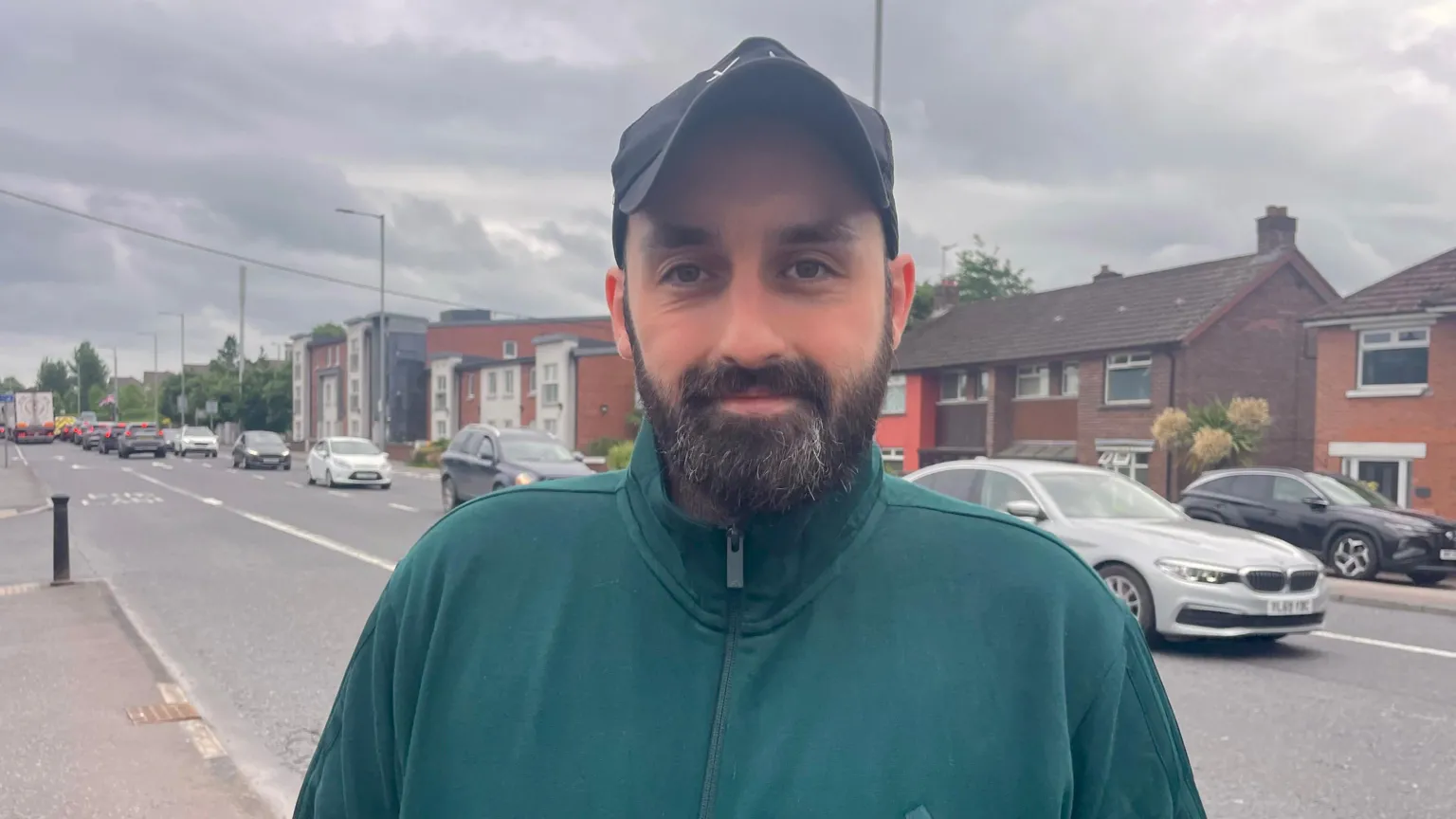 A man, with brown hair and a brown beard, wearing a dark green top and a black cap. He is standing on the side of a road with a row of houses behind him. 
