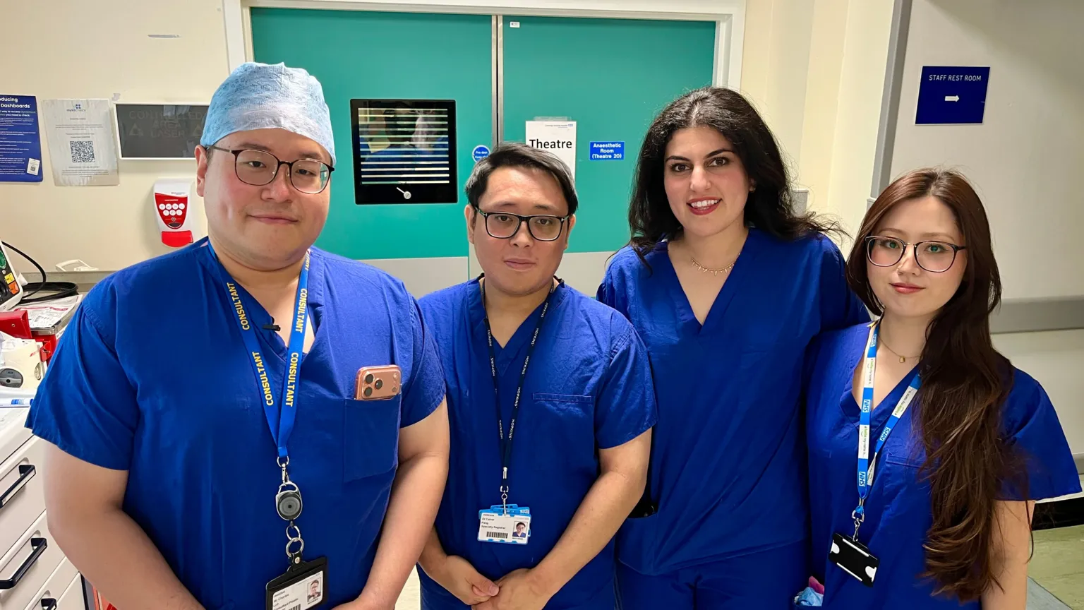 MARTIN GILES/BBC A team of clinicians in blue scrubs. Three have lanyards around their neck. They are standing in front of the doors to one of the theatres