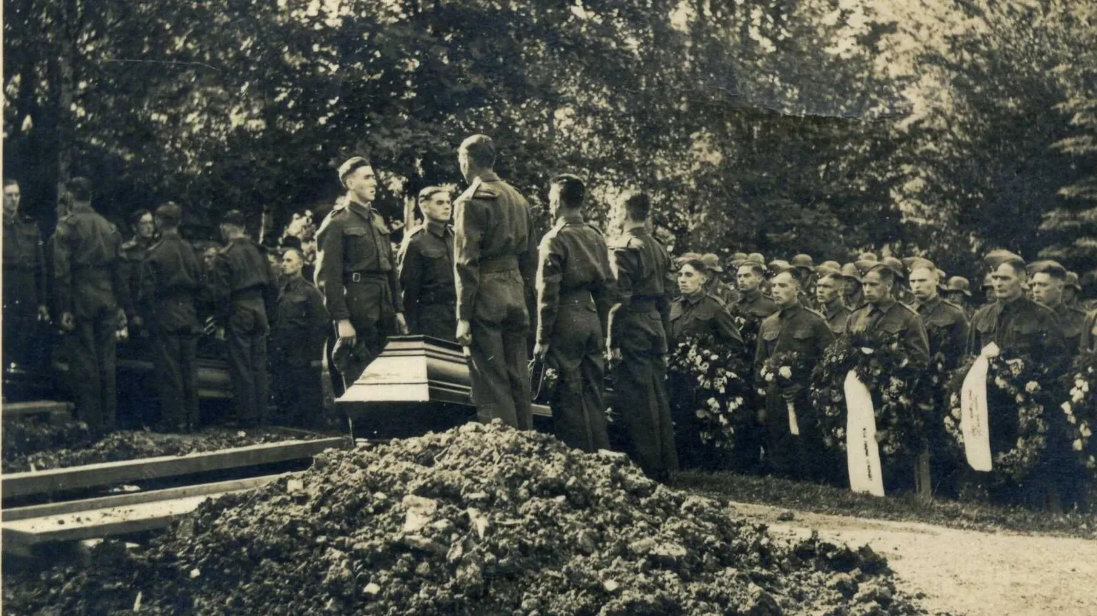 Libby MacRae A monochrome photograph from the 1940s which shows a burial ceremony in Berlin. In the foreground there is a pile of earth which has been removed from a freshly dug grave. Five soldiers in uniform can be seen standing to attention by a wooden casket. Further ranks of soldiers holding ceremonial wreaths stand in the background. 