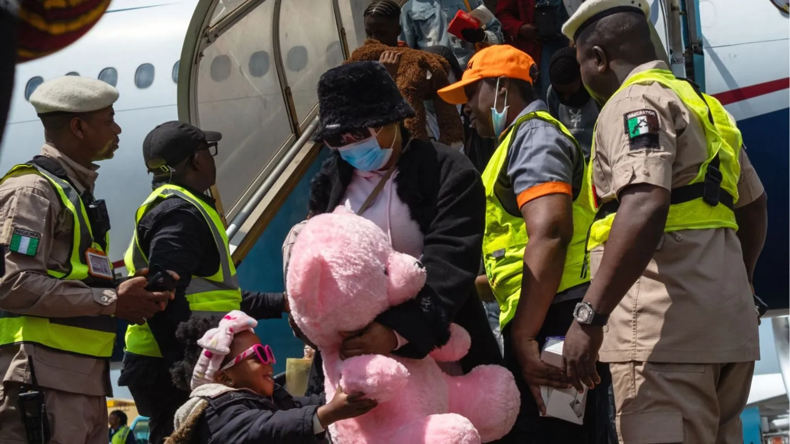 AFP via A young girl in pink sunglasses holds a giant pink teddy as she and a woman disembark from a flight in Nigeria - security officers seen either side of them