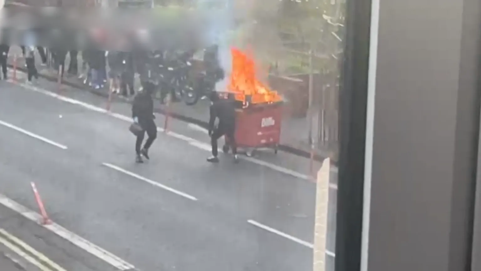 Tejal Samat and Nishath Mohammed Picture take from window showing two people in dark clothing setting fire to a bin. A crowd of people are standing on the footpath.