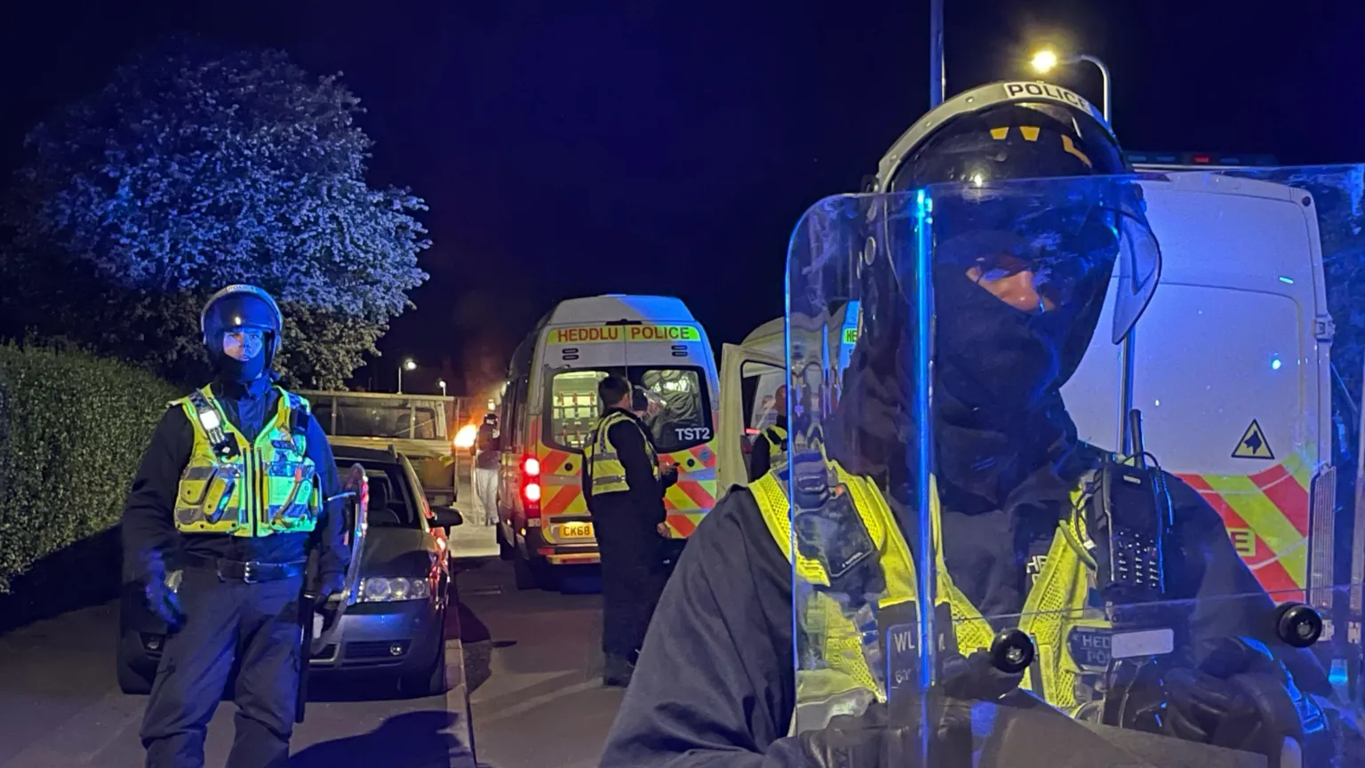 PA Police officers shown on an Ely street holding riot shields and wearing police helmets. Two police vans can be seen in the background.