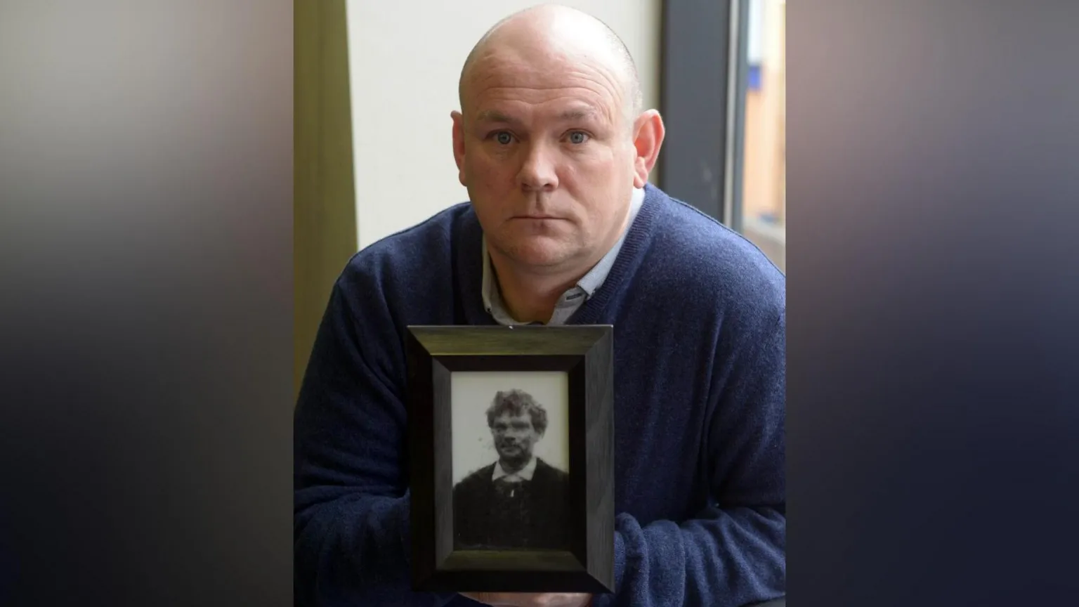 Pacemaker Nephew of John Pat Cunningham at a press conference in Belfast after his family received an apology from the government. He is bald, wearing a blue jumper with shirt underneath, holding a framed picture of John Patrick Cunningham.