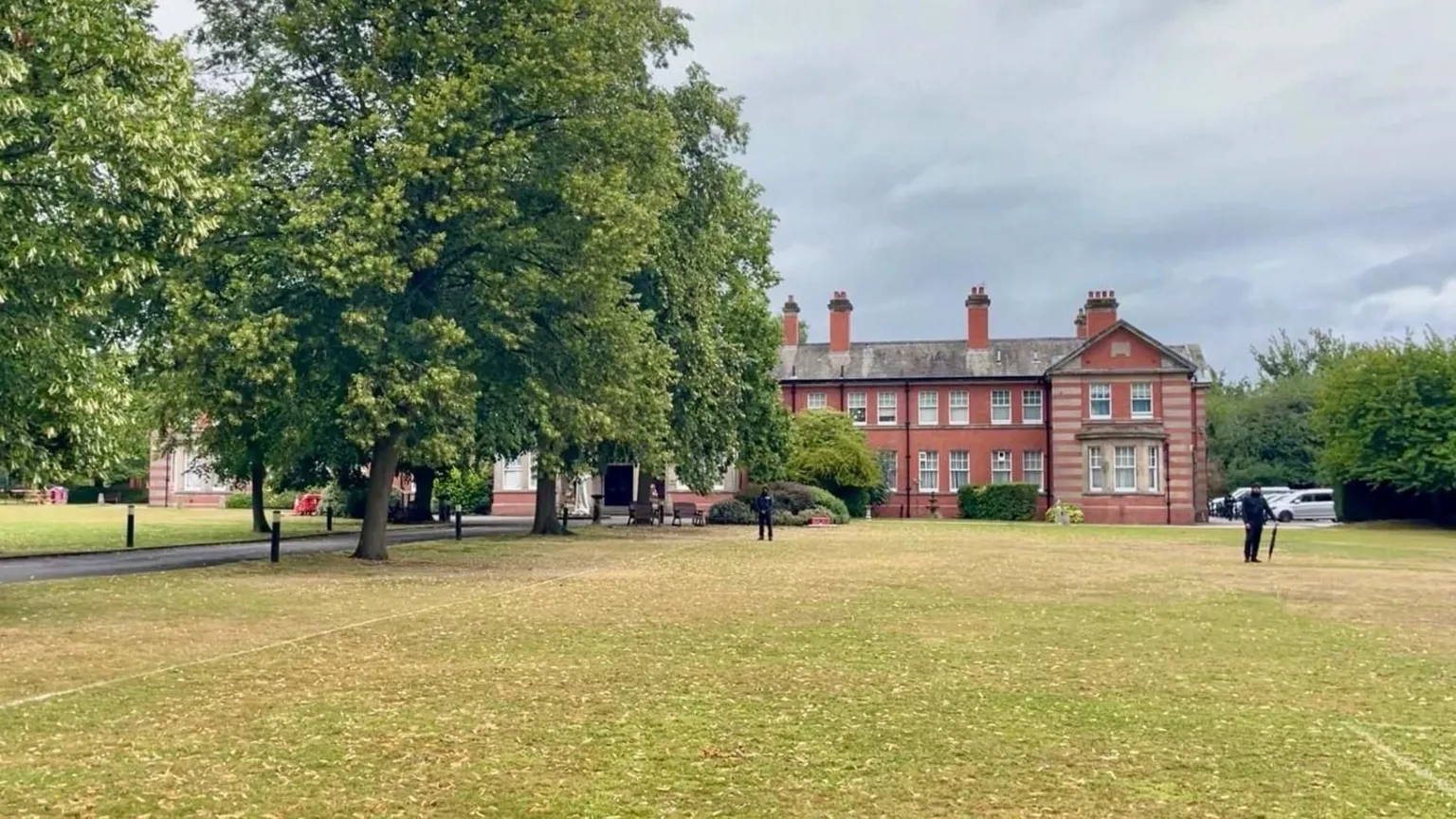 An ornate building with a large field in front of it as well as a playing field and trees. There is a row of trees to the left hand side of the image, obscuring part of the building. Two people in black clothing are standing in front of the building.