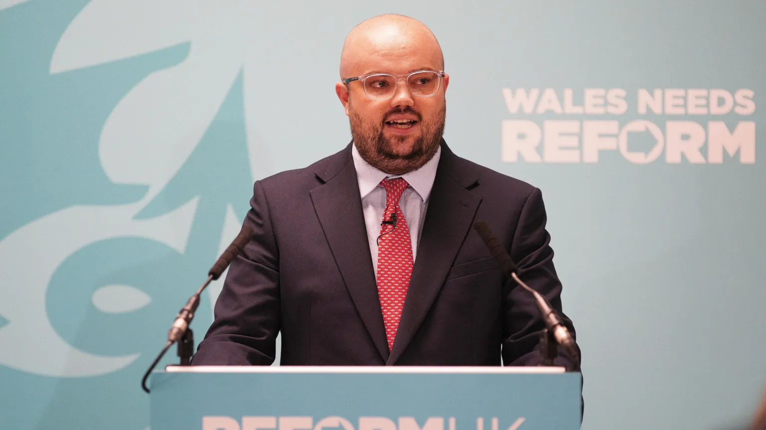 Llyr Powell speaking at a teal lectern which reads