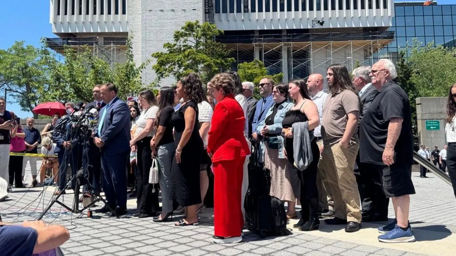 At a distance, a man stands at a mic with a group of people surrounding him. they are in front oiif a courthouse outside with blue sky behind them