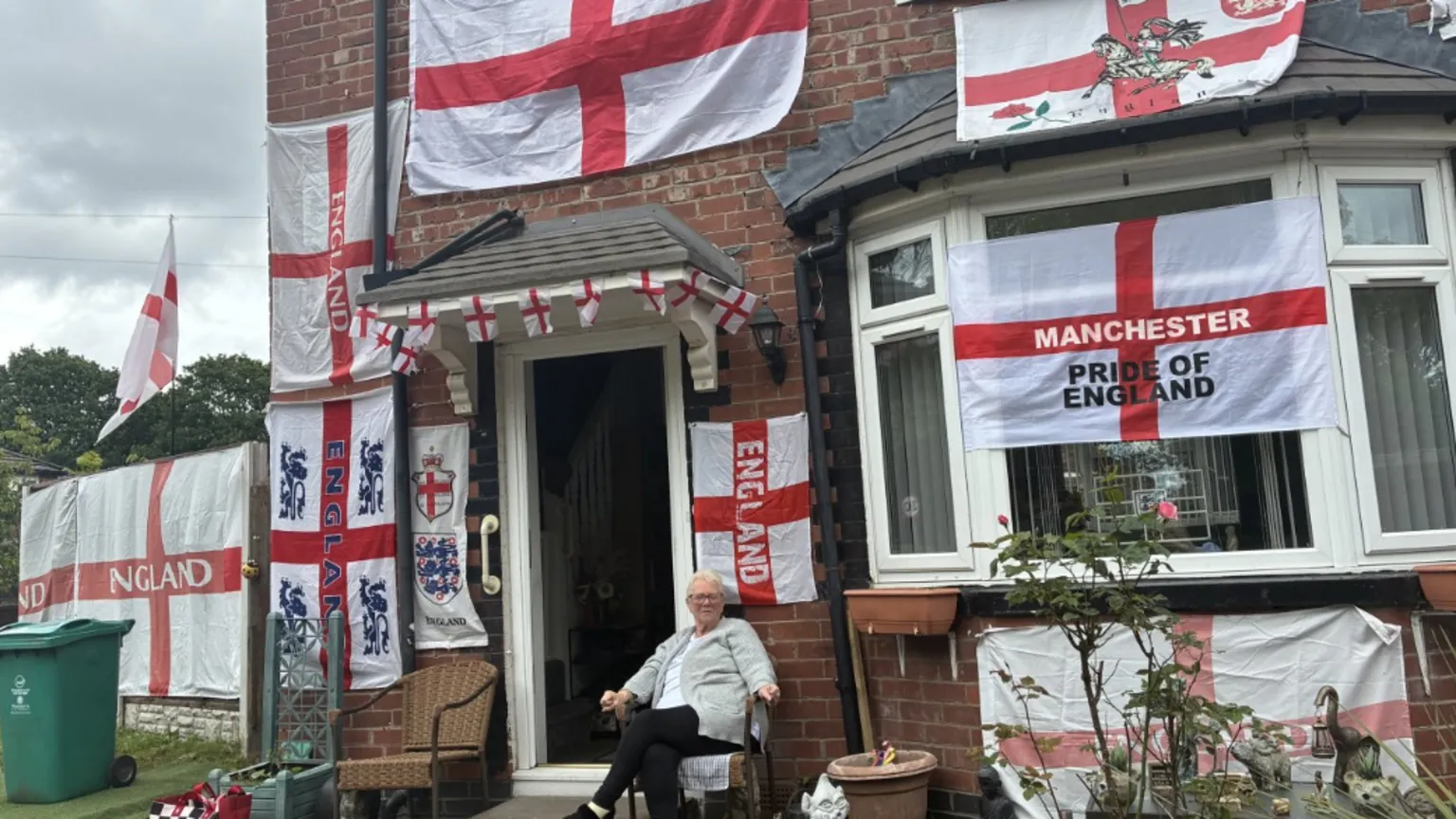 Mary, a woman with short white hair and glasses, sits in front of a house covered with various England flags.