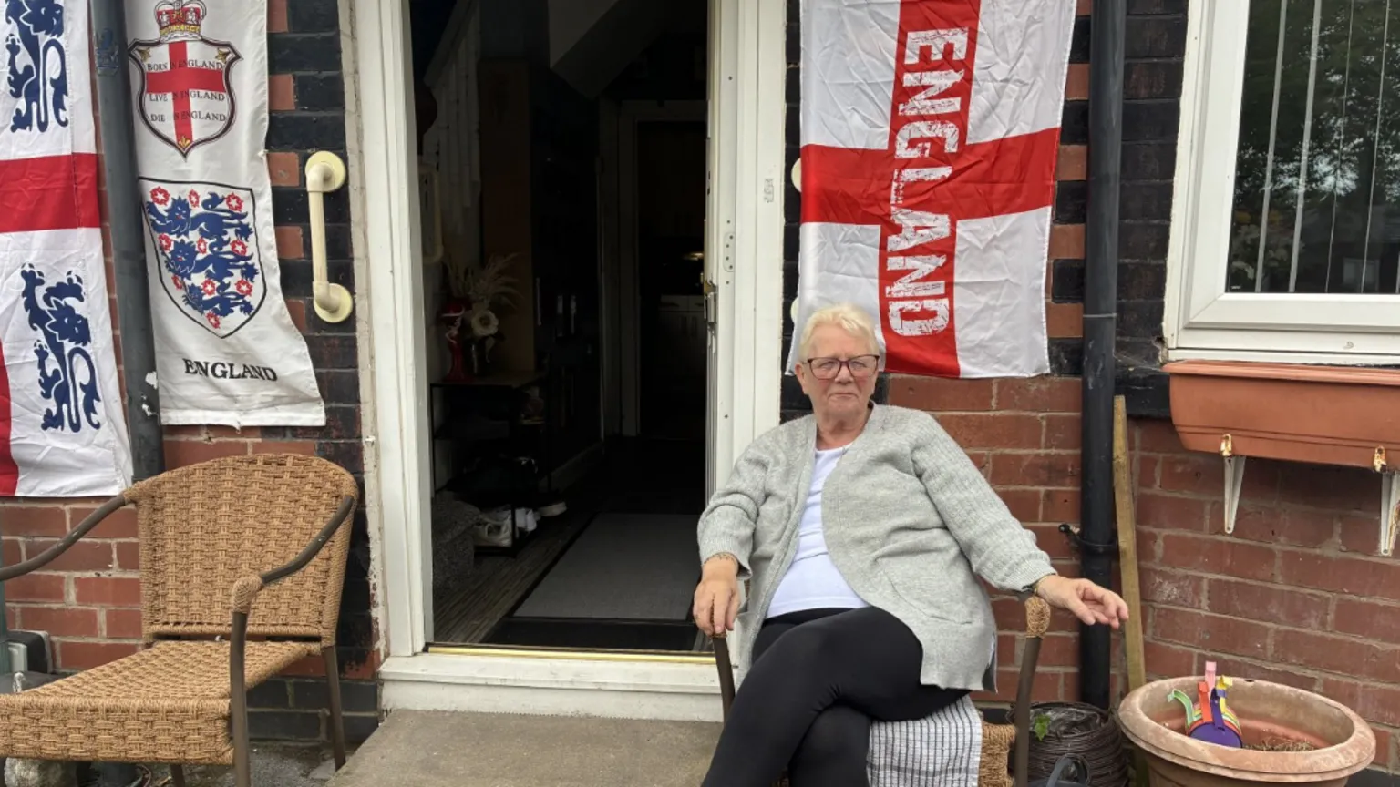 Mary, a woman with short white hair and glasses, sits on a chair in front of a house covered with various England flags.