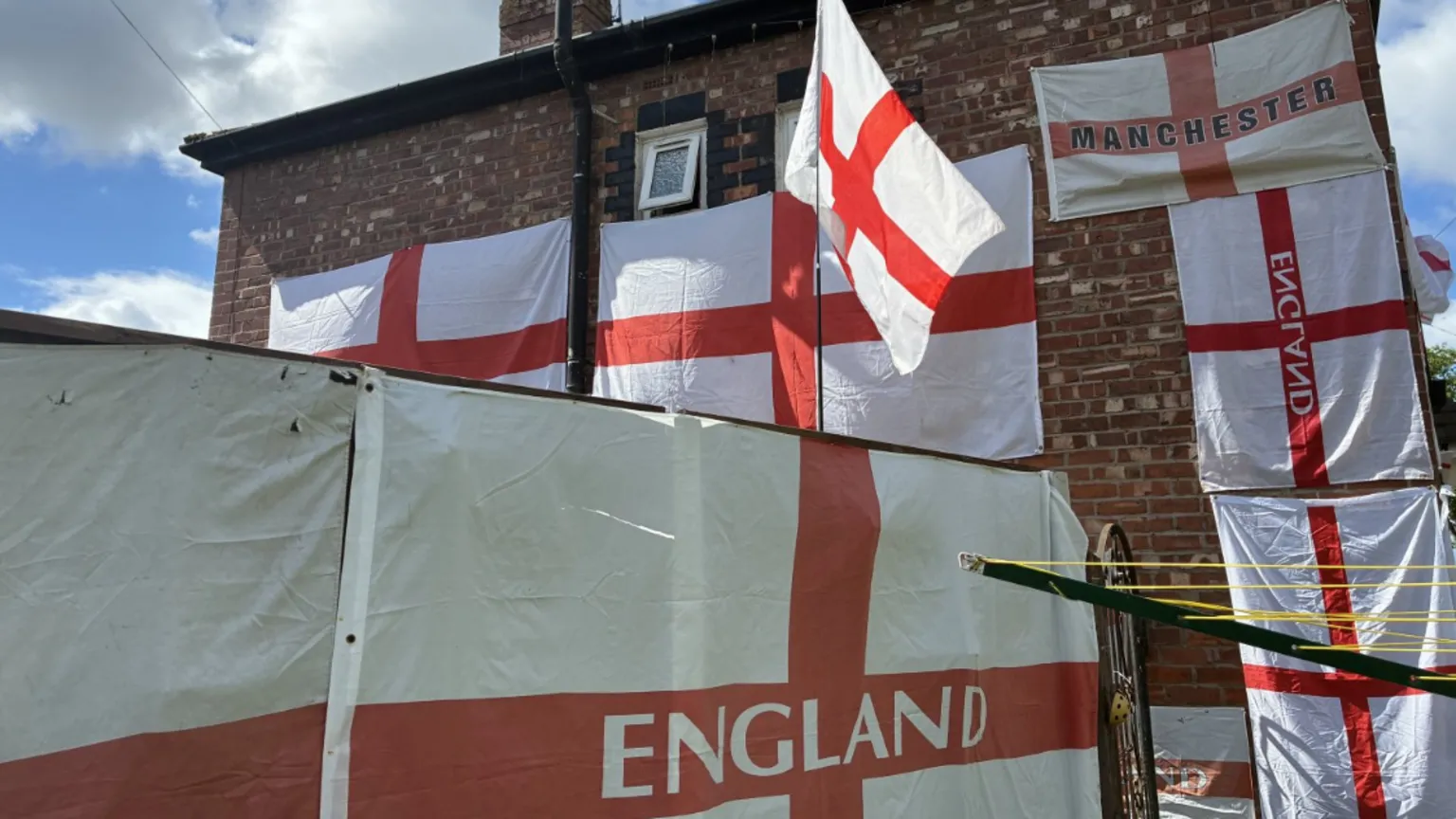 The side wall of a house covered with many England flags with one flag flying on a flagpole atop a wooden garden fence.
