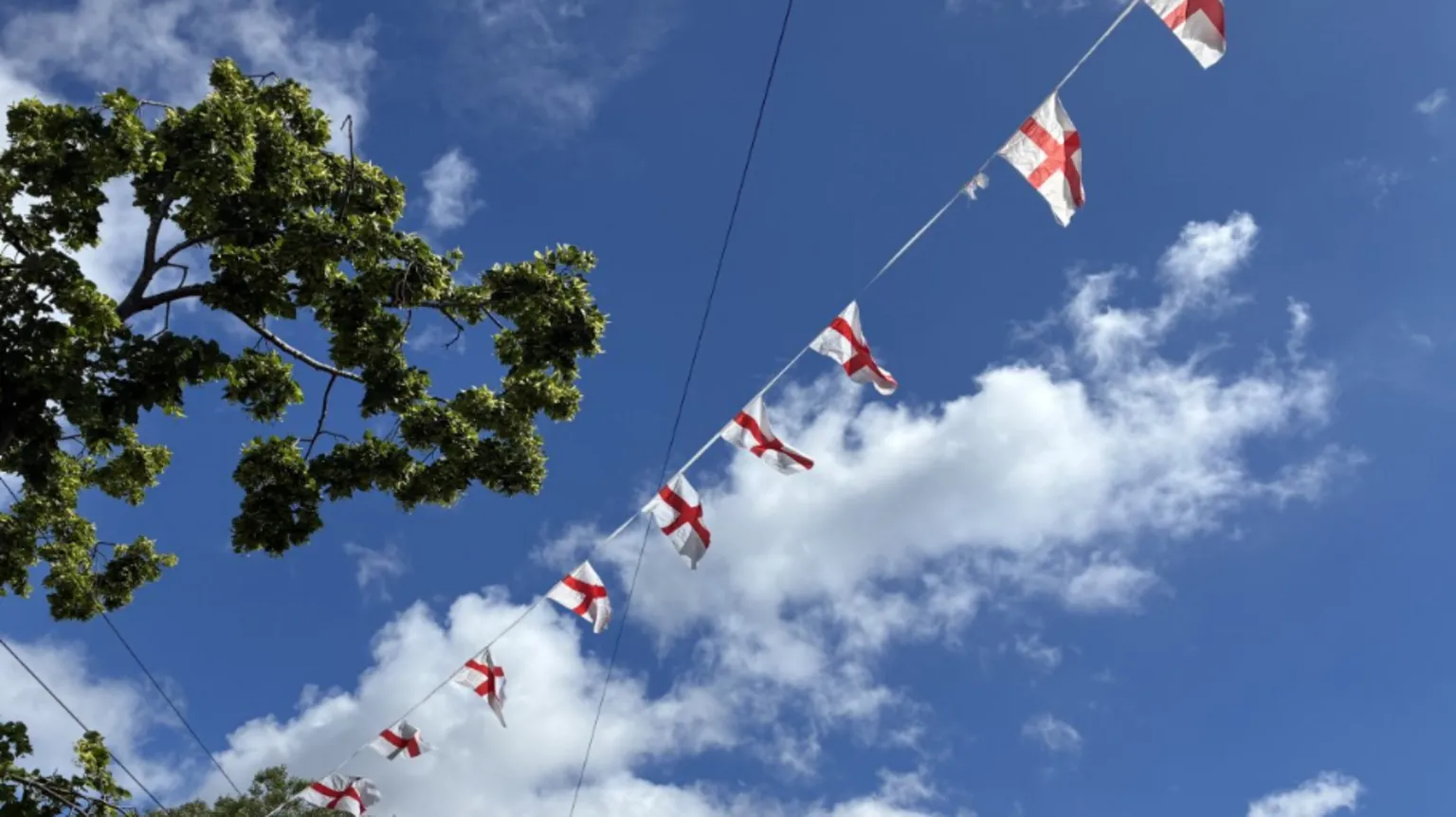 England flag bunting flies in a blue sky with a couple of clouds. A green leafy tree is visible in the top left of the picture.