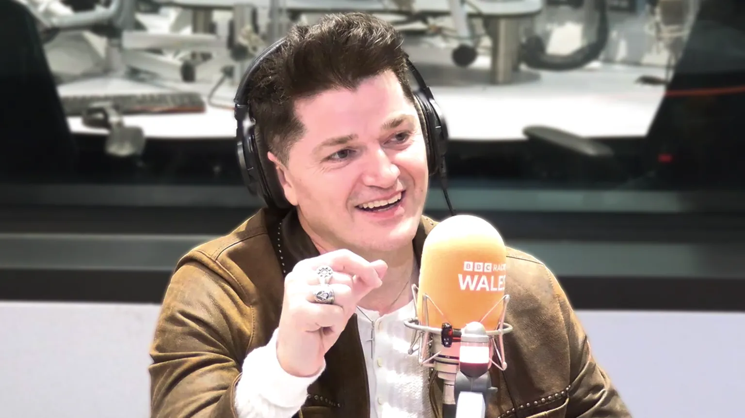 Danny O'Donoghue in a BBC Radio Wales studio. He is sat in front of an orange BBC Radio Wales microphone and is wearing black headphones. He is wearing a green jacket and a white long sleeve top. He is wearing two large silver rings.