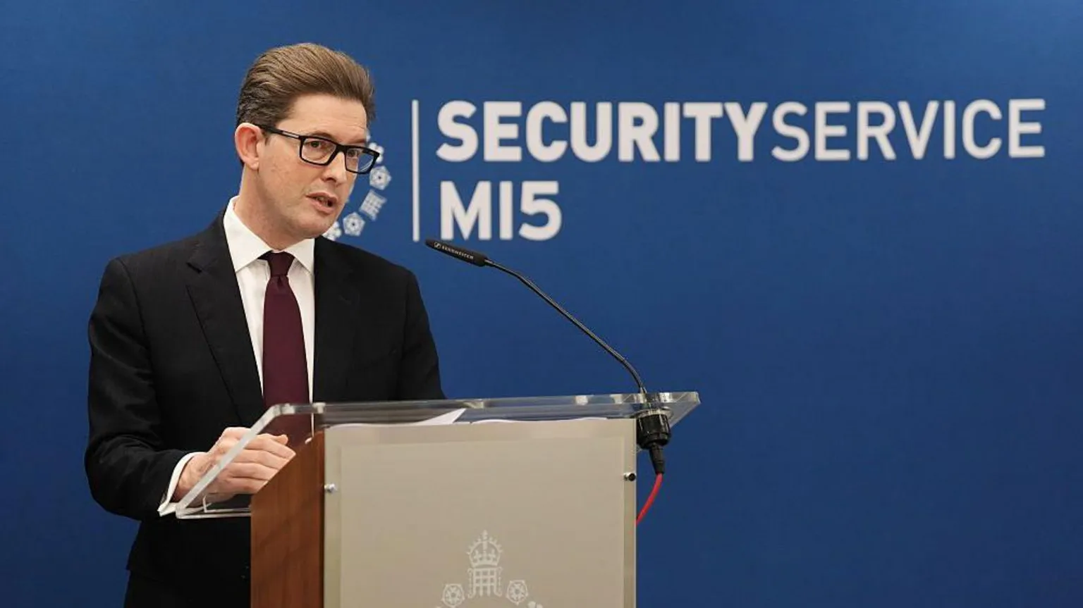 Sir Ken McCallum standing behind a podium with the MI5 insignia emblazoned on the front in front of blue background with the words 'MI5 security service' behind him. Sit Ken is wearing a black suit, a dark maroon tie, white shirt, black-framed glasses and clicked back brown hair.