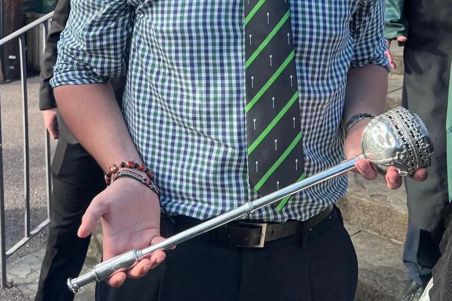 Caleb Tutt A silver mace being held by a man. All you can see is the mace and the hands of the man, as well as his torso. He is wearing a checkered white, purple and green top with a striped dark green and light green tie.