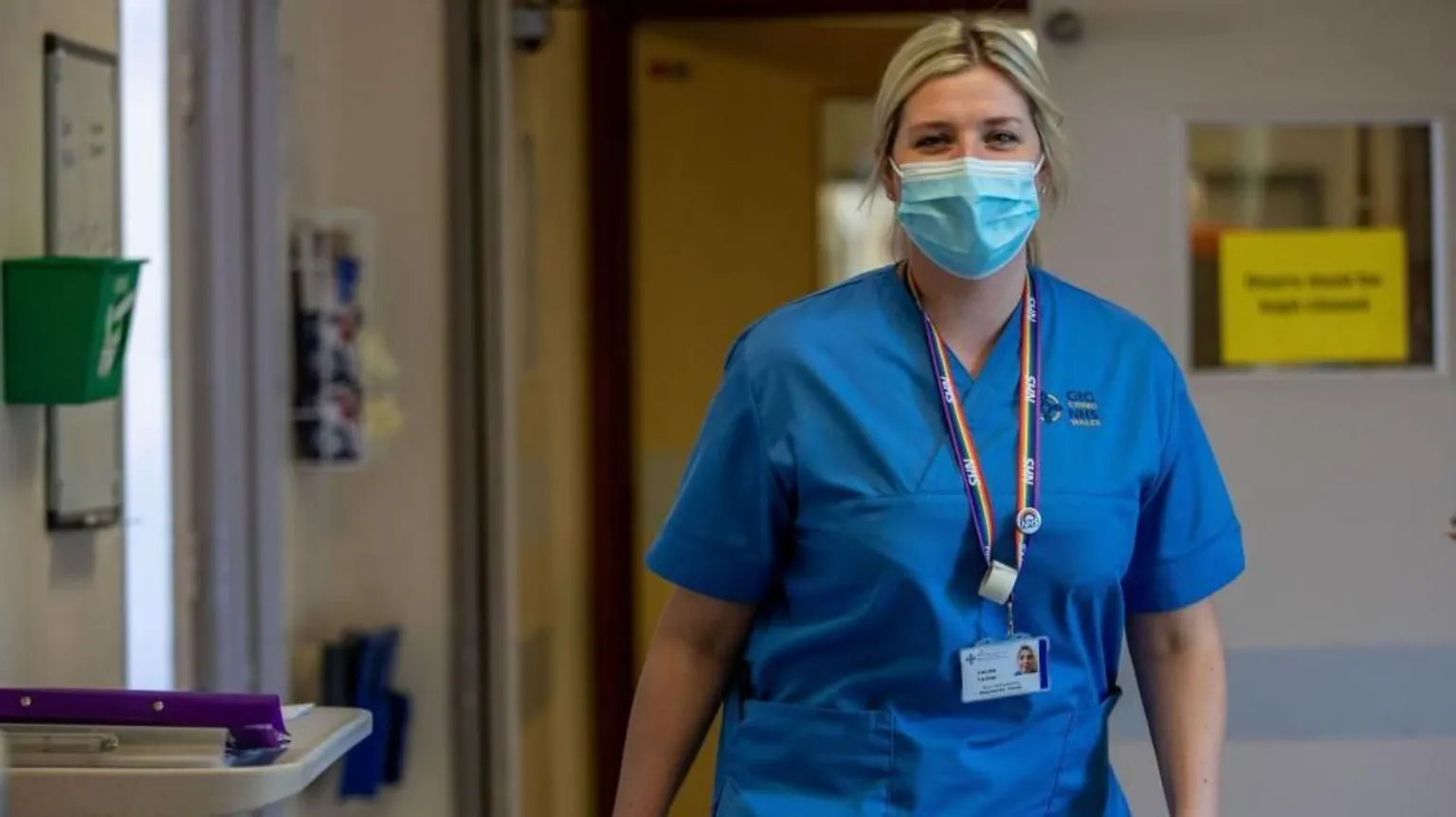 A nurse wearing face mask in blue scrubs, a blue face mask and a rainbow lanyard walking down a ward