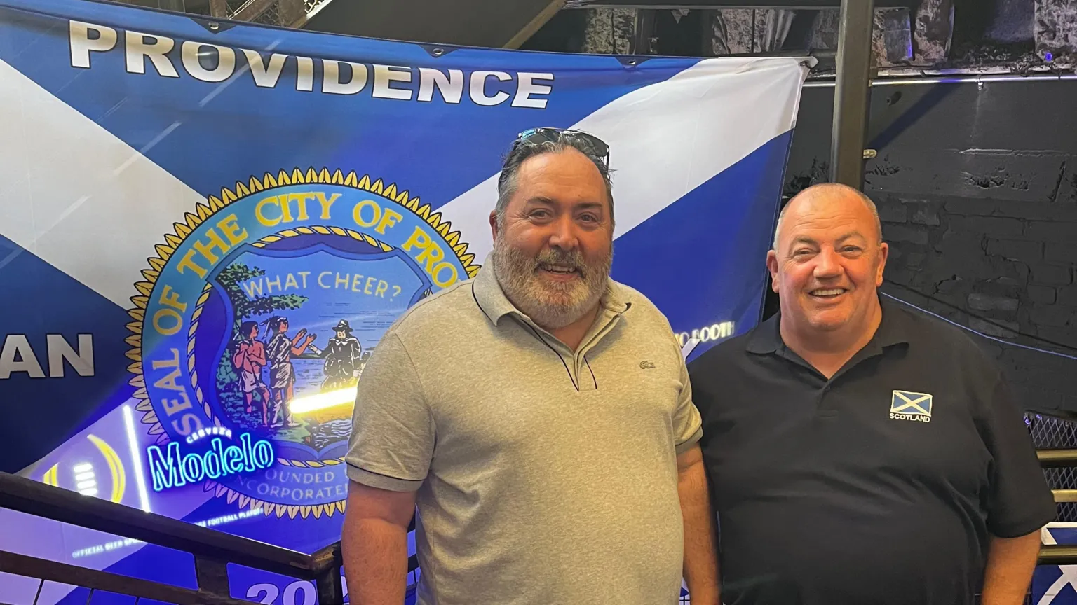 A man with dark hair and dark facial hair wearing all grey, standing next to a smaller bald man who is wearing navy with a saltire on his polo shirt, they are standing in front of a saltire with the world providence written on it. 
