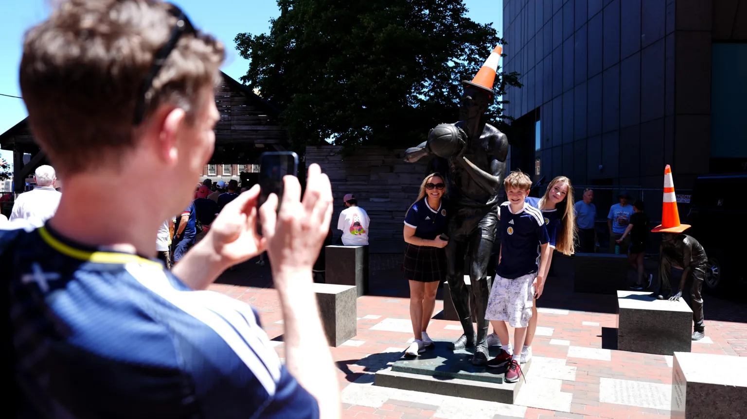 A man taking a photograph of two women and a boy wearing blue Scotland football shirts standing next to statues with orange traffic cones on their heads.