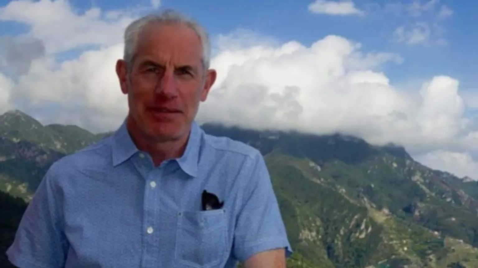 Family photo Tudor, with grey hair in a blue short sleeve shirt. He's standing in front of a mountain range and blue sky and clouds are seen in the background.