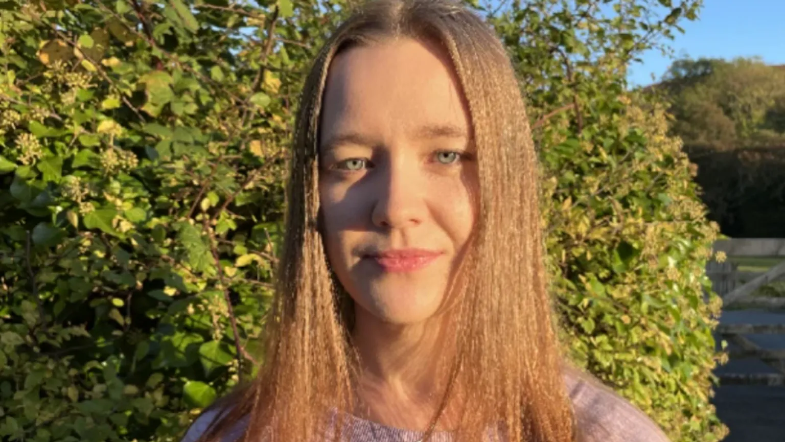 Bethan, with long blonde hair, wearing a pink t-shirt and standing in front of a green hedge. She is looking directly at the camera with a neutral expression.