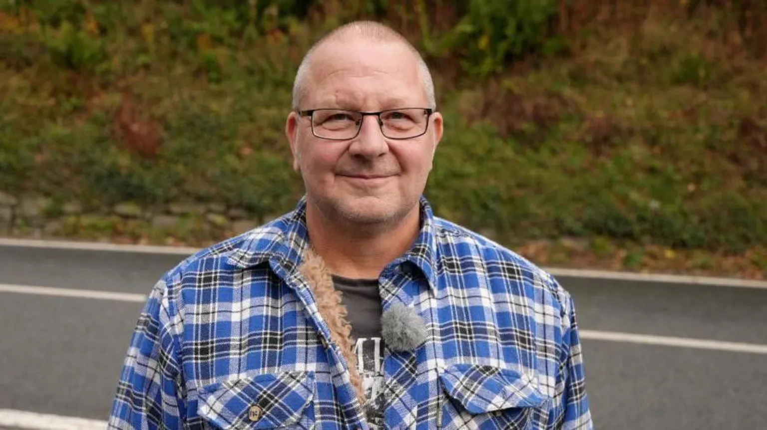A bald man with glasses wearing a black and blue check jacket. He is stood in front of a road and is looking at the camera with a small smile.