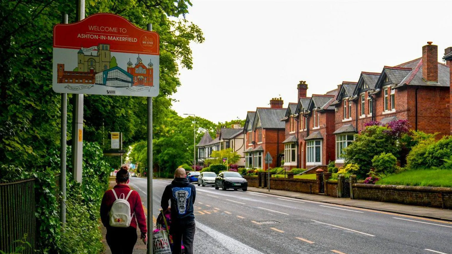 Two people walk under a sign, one pushing a pram, along a roadside in Ashton-in-Makerfield, Greater Manchester.