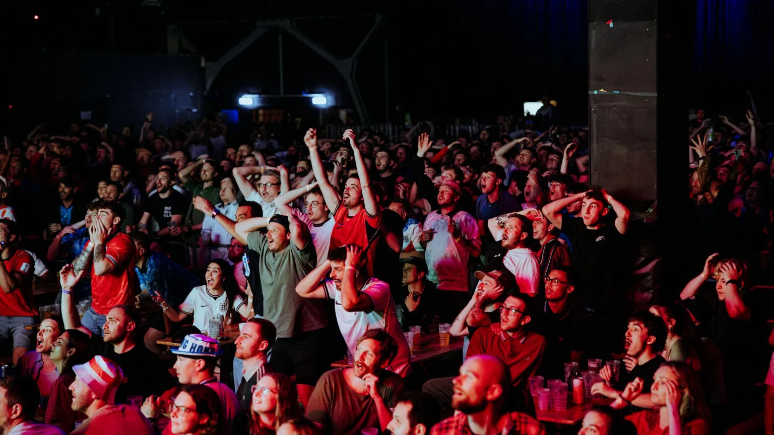 Alastair Brookes A large crowd of England football fans at a watch party in Bristol. Men and women can be seen and many are wearing football tops and other merch like hats. Many of the fans have their arms up in the air or on their head with their mouths open.