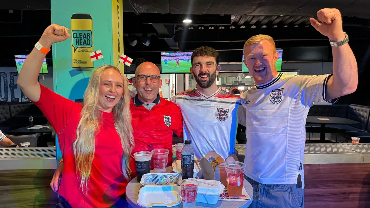 Three male and one female England football fans at a watch party in Ashton Gate Stadium in Bristol. They are all wearing England football tops and the woman has a headband with two England flags on it. They are stood at a table full of drinks and food, huddled, with their arms up and smiles on their faces.