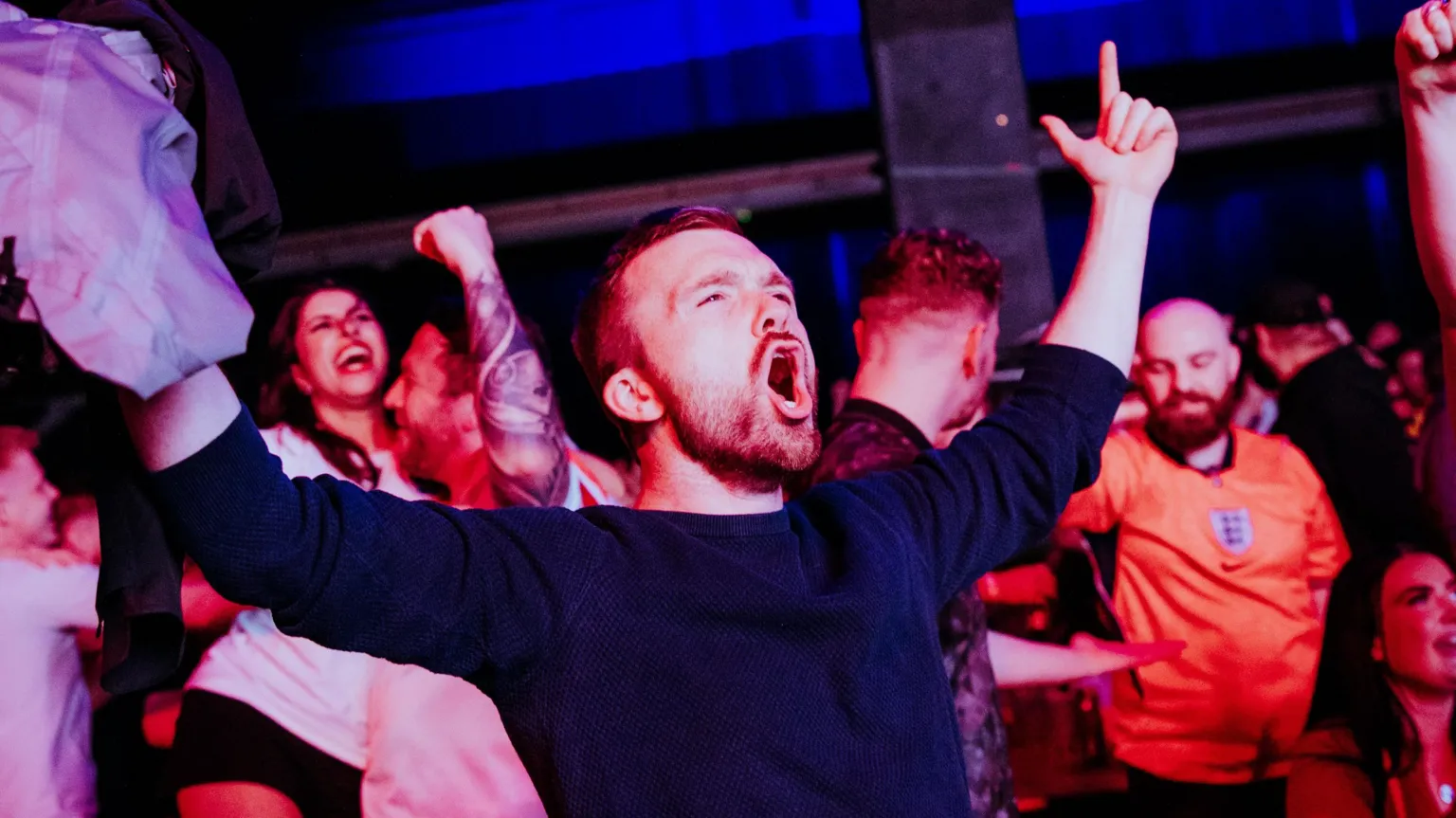 Alastair Brookes A man celebrating England's success in a World Cup match. He has dark short hair and facial hair and is wearing a dark coloured top. His mouth is open and he is pointing upwards.