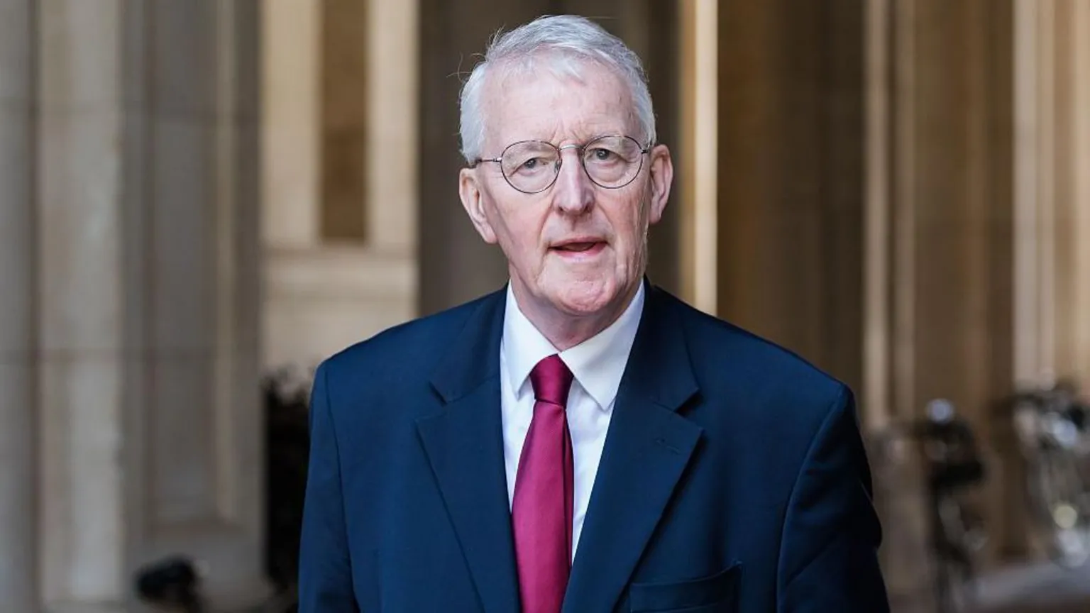 Hilary Benn speaking. He is wearing a navy suit jacket, white shirt and red tie. He has grey hair and is wearing round glasses.