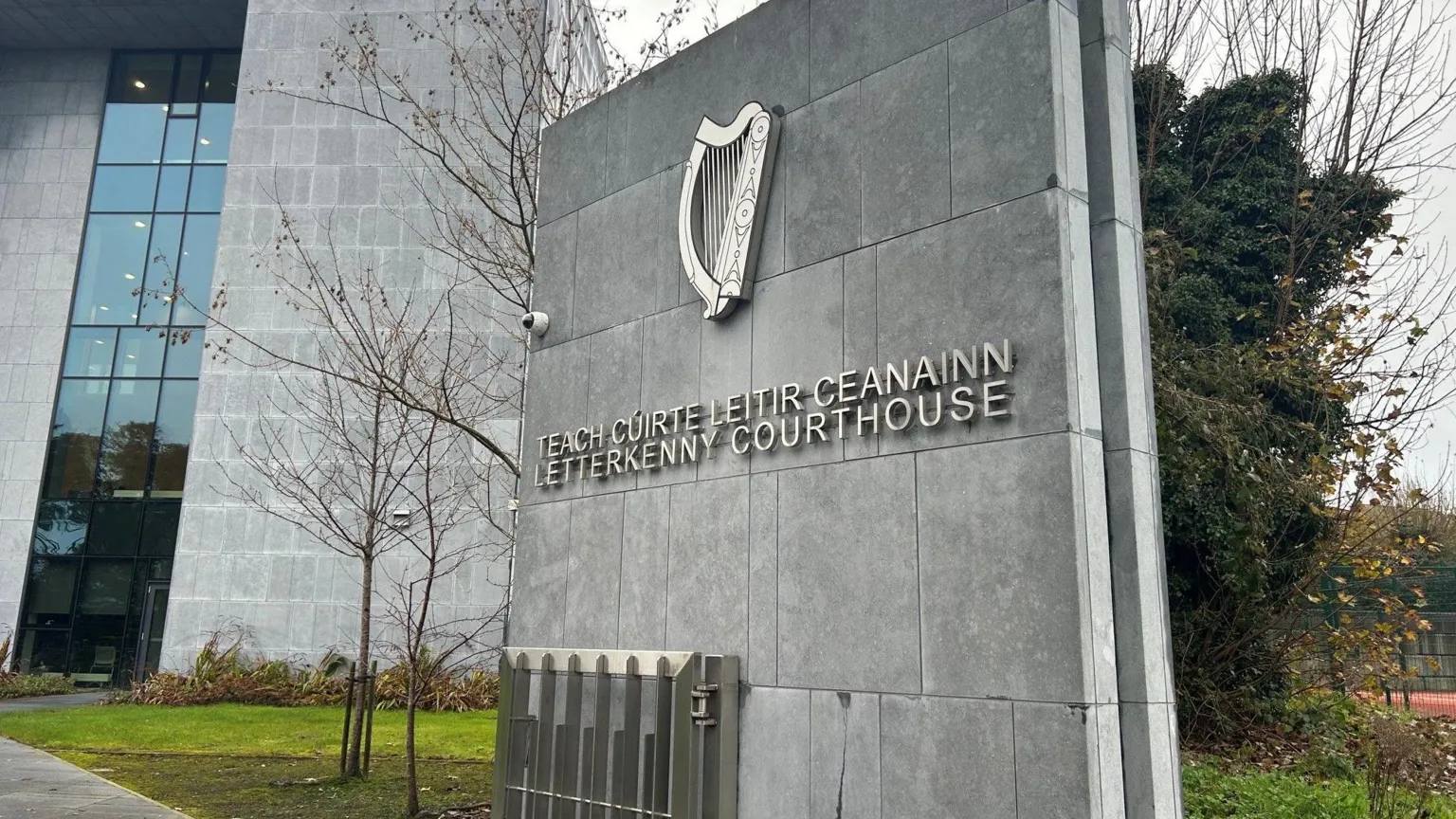 A large grey stone wall stands in a garden area outside a larger building made of the same material. It has a large stainless steel harp at the top followed by two rows of writing. The top line is in Irish reading 'Teach Cúirt Leitir Ceanainn'. Below is the English translation 'Letterkenny courthouse'.