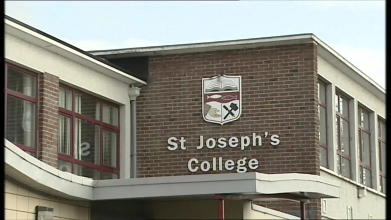A large school crest is hung on the side of an old red-bricked building. Underneath it is a sign which reads 'St Joseph's College' in white text. The crest depicts a book, the outline of two fish facing each other, a hand reaching out open palmed and a hammer and chisel crossed over each other.