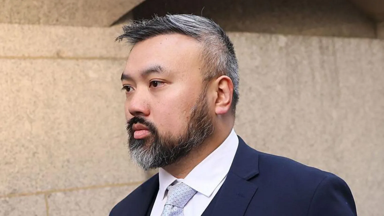 A shot of Chi Leung “Peter” Wai from the shoulders up. He has black and grey hair and a black and grey beard. He is wearing a navy suit with a white shirt and a light blue tie. He is walking and looking to the left.