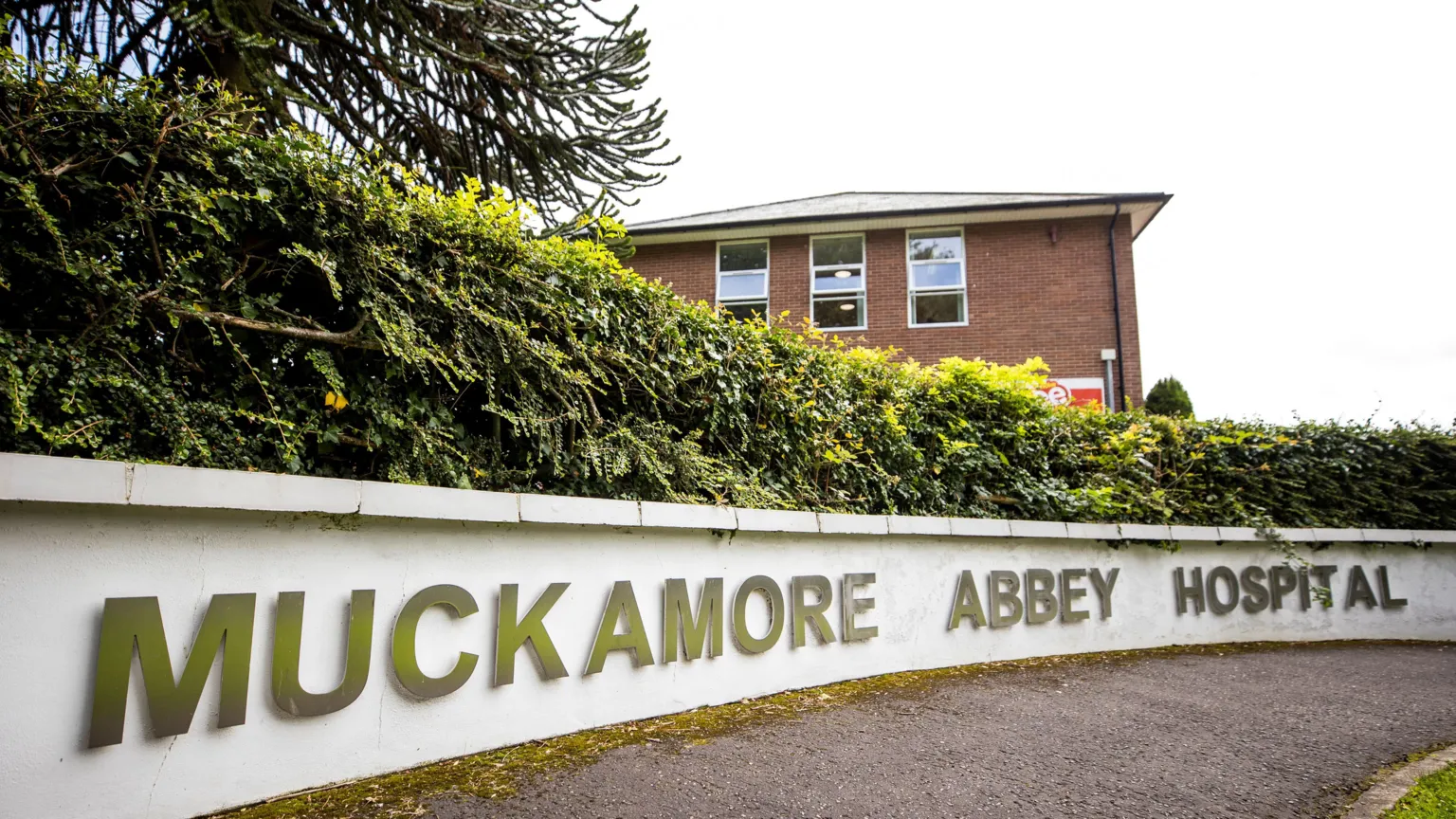 Muckamore Abbey Hospital - the name is in big capital letters along a low curved white wall. There is a path and patch of grass in front. There is a hedgerow behind it and then behind that a red brick building with three of the top windows visible behind.