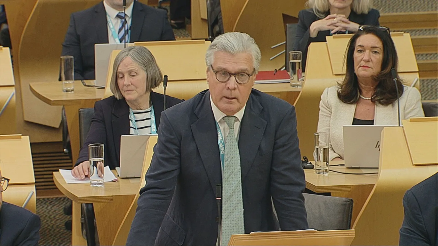 Scottish Parliament Malcolm Offord, wearing a dark suit and green tie, stands to speak in the Scottish Parliament