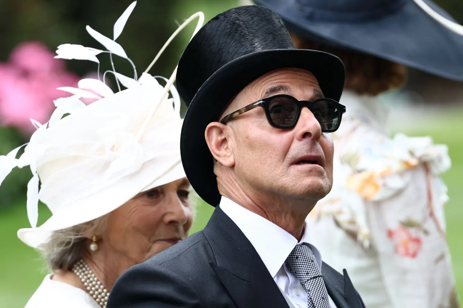 Actor Stanley Tucci looking up at something at Royal Ascot. He is wearing sunglasses and is wearing a suit.