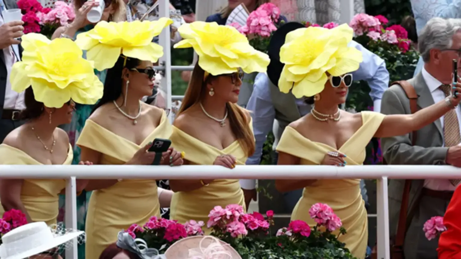 Four women, wearing yellow hats that look like flowers, lined up, with the woman on the right hand side taking a selfie of them on a mobile phone.