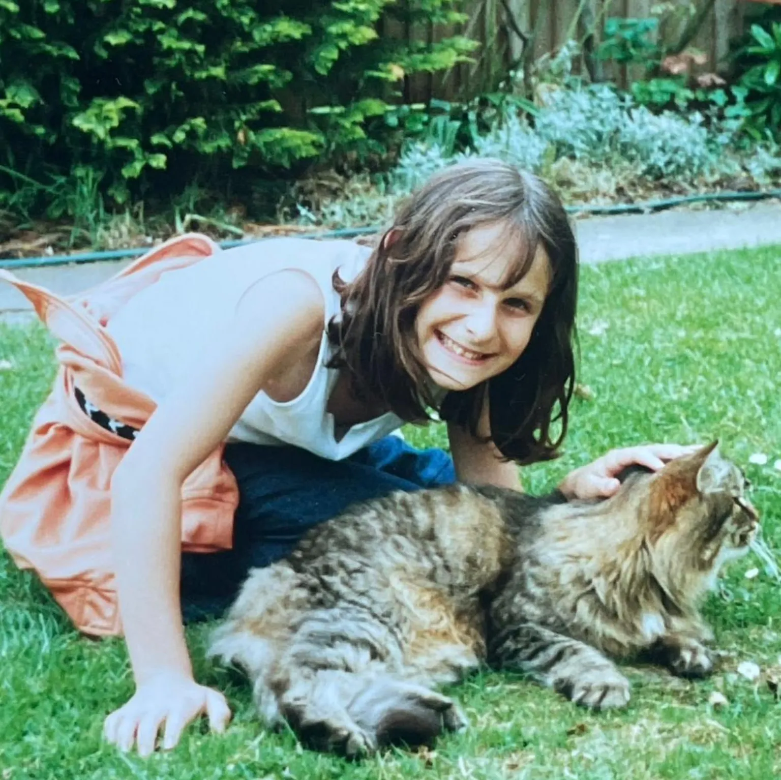 Natasha Goldstein-Opasiak A young girl in the garden with a long haired tabby cat. She's smiling at the camera and has long brown hair. She is wearing blue jeans and a white, sleeveless top with an orange tracksuit top tied around her waist.
