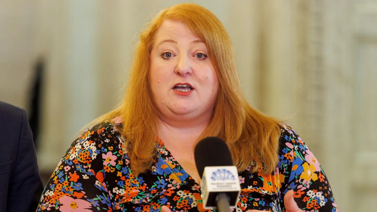 Naomi Long speaking into a microphone. She is wearing a patterned top and has long red hair.