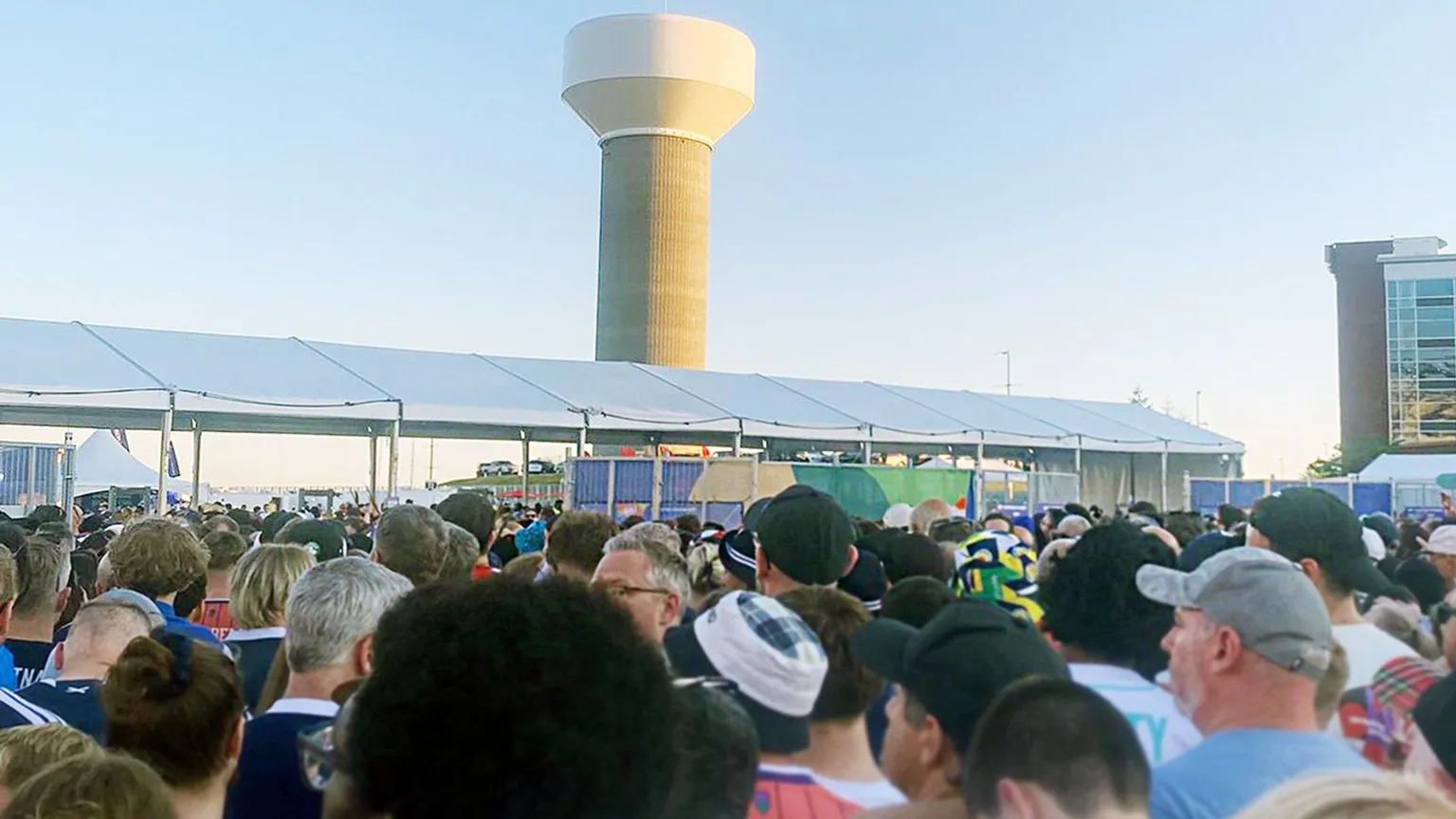 Age crowd of football fans, viewed from behind, massed outside a stadium at a security checkpoint.