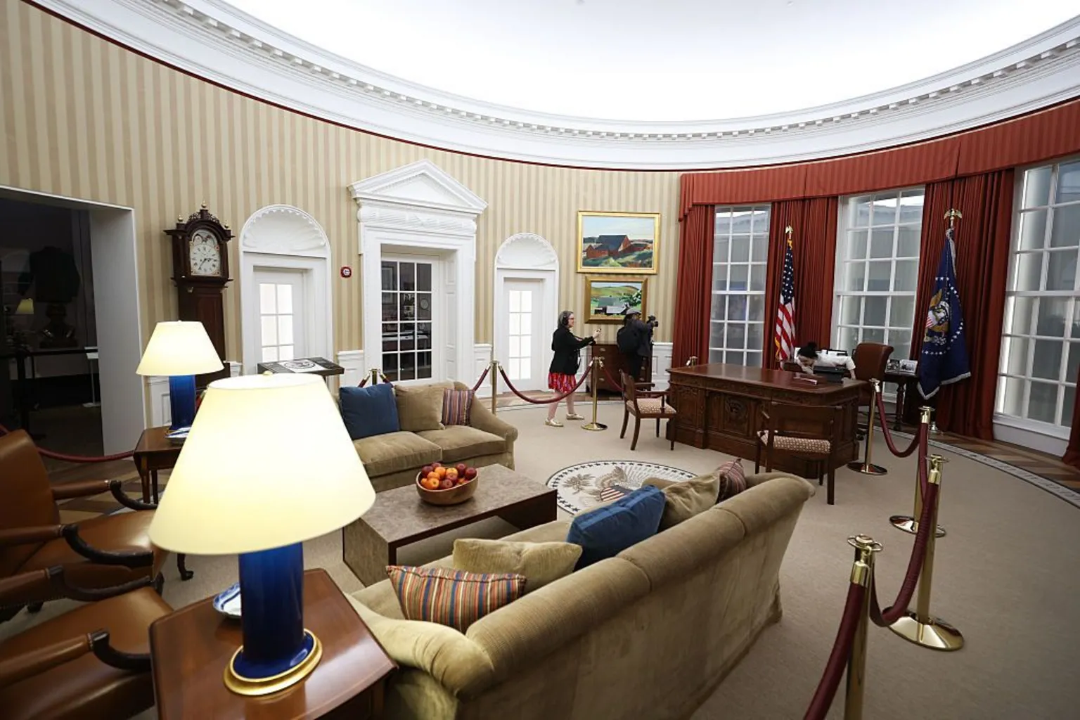 Talia Sprague/Bloomberg via Stuffed chairs, lamps and dark wood furniture fill a replica of the Oval Office, with parts of the space sectioned off by red velvet ropes. Two people example the space.