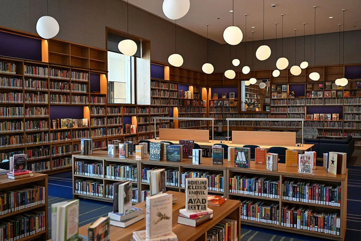 Joshua Lott for The Washington Post via A room is lined with bookshelves and filled with book displayed made of light wood. There is a blue and green carpet and irregular orb lights hang from the ceiling.
