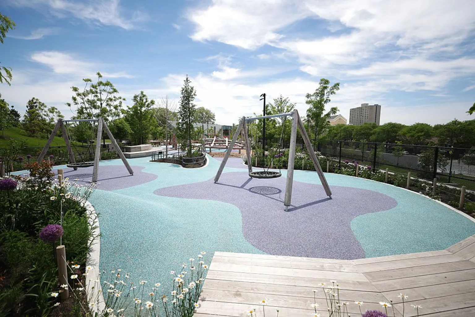 Talia Sprague/Bloomberg via Two sets of swings are shown on a teal and purple playground surface. They are surrounded by lush landscaping and wooden benches and walking paths.